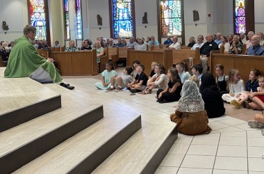 A priest sits on the floor with children and speaks to them during his homily at Mass