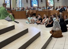 A priest sits on the floor with children and speaks to them during his homily at Mass