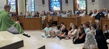 A priest sits on the floor with children and speaks to them during his homily at Mass
