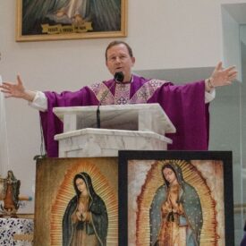 A priest dressed in a purple vestment spreads his arms open wide in a welcoming gesture during a homily