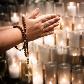 A woman's hands folded in prayer on All Souls Day in front of rows of lit candles