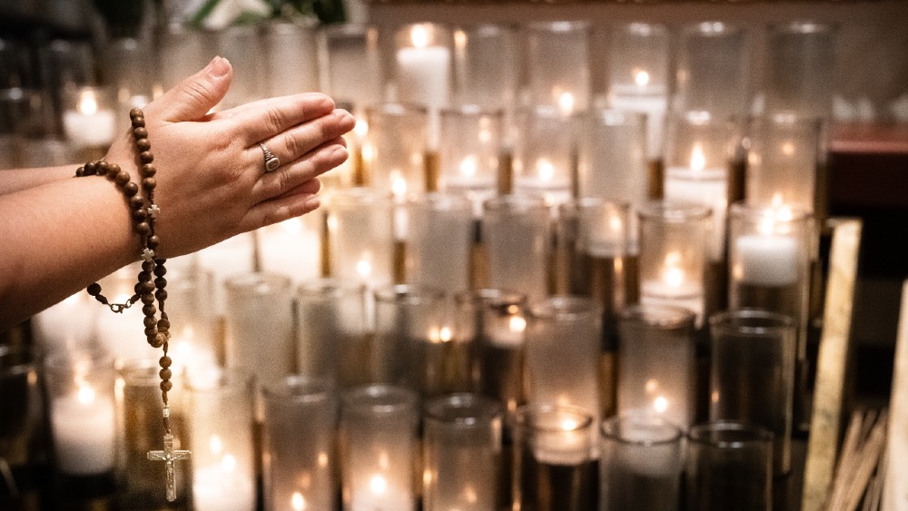 A woman's hands folded in prayer on All Souls Day. She prays in front of rows of lit candles with a rosary wrapped around her wrists.