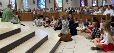 A priest sits on the floor with children and speaks to them during his homily at Mass