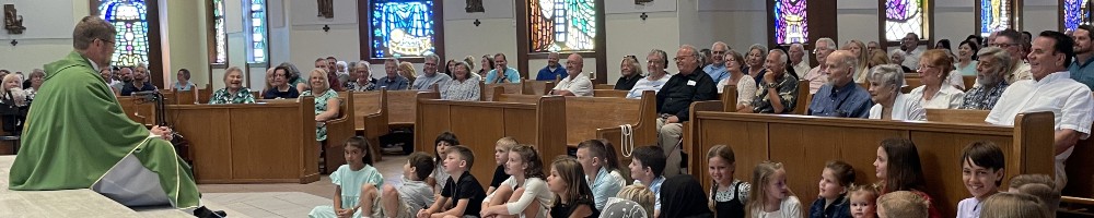 A priest sits on the floor with children and speaks to them during his homily at Mass