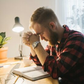 A man sitting at a desk with his hands folded and head bowed in prayer